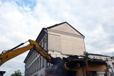 Manipülatörlü bir inşaat kazıcısı, bulutlu mavi bir gökyüzünün arka planına karşı eski bir evi söker ve yok eder. İnşaat alanı ve ağır ekipman