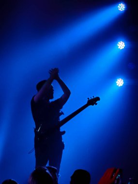 Müzisyen gitarist sahnede mavi spot ışıklarıyla gitar solosu yapıyor. Kimliği belirsiz bir kişi ve bir müzik olayı