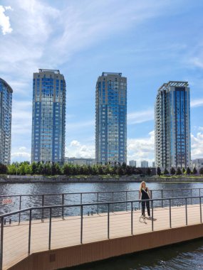 Genç bir kadın, Jack Russell teriyeri köpeğiyle köprüde yürüyor ve gülümsüyor. Arka planda şehir gökdelenleri var