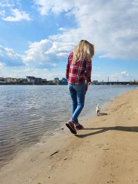 Sarışın bir kız nehir kıyısındaki sahilde Jack Russell teriyer köpeği perspektifinde yürüyor. Arka görünüm