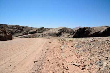 Açık mavi gökyüzünün altındaki tepelere uzanan bir çöl bölgesinde toprak bir yol.