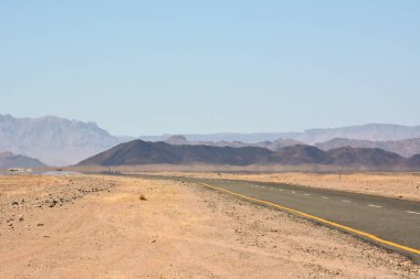 Çölde düz bir yol, perspektif olarak, açık mavi gökyüzünün altındaki dağlara çıkıyor.