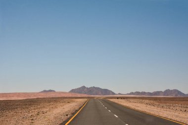 Çölde düz bir yol, perspektif olarak, açık mavi gökyüzünün altındaki dağlara çıkıyor.