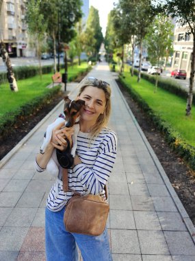 Sarışın bir kız gülümser ve Jack Russell Terrier cinsinden bir köpeği kollarında tutar ve şehrin bulvarında perspektif olarak yürür. Arkaplan bulanık