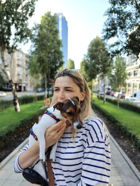 Sarışın bir kız gülümser ve Jack Russell Terrier cinsinden bir köpeği kollarında tutar ve şehrin bulvarında perspektif olarak yürür. Arkaplan bulanık