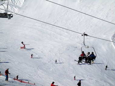 Kayakçılar kayak merkezindeki karlı bir dağ yamacının arka planında kayak asansörüne binerler. Kış sporları