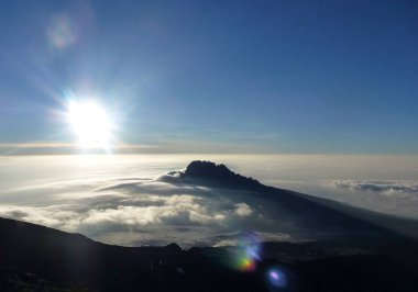 Bulutların üzerinde yükselen yakın bir dağın üst görüntüsü parlak güneş ve mavi gökyüzünün arka planına karşı. Resimli dağ manzarası