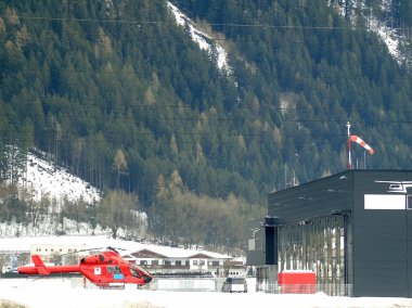 A red helicopter of the rescue service stands near the hangar on the background of a winter mountain covered with trees in a dense forest clipart