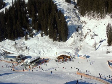 Karla kaplı kayak yamacı ve dağ manzarası. Aşağıda turistlerin bulunduğu kayak merkezinin binaları da yer alıyor.