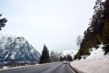 View of a picturesque asphalt road between a snowy mountain and a forest. A car is driving ahead in perspective. Winter tourist landscape clipart