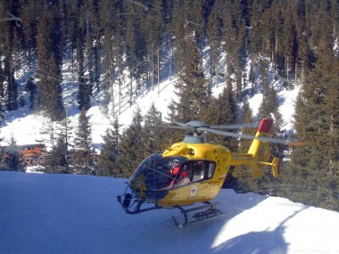 Sarı kurtarma helikopteri bir kayak merkezinin yamacından kalkıyor. Arka planda ağaçlardan oluşan bir orman var. Kurtarma ekipmanları ve doğa