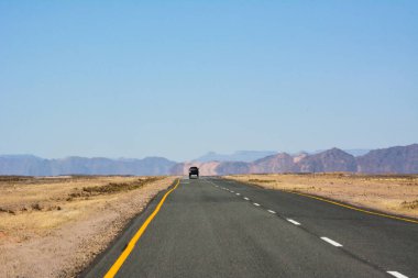Çölde düz bir yol, perspektifte bir de uzaktan giden bir araba. Turist seyahati. Uzaktaki küçük dağlar