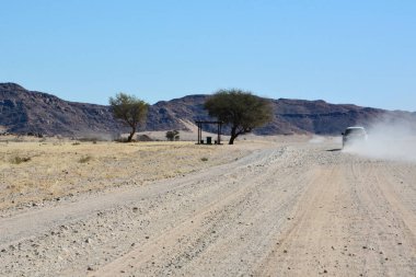 Bir araba çölde toprak bir yol boyunca tepelere doğru perspektifle ilerliyor. Arabanın arkasından toz yükseldi. Turist rotaları ve seyahatleri