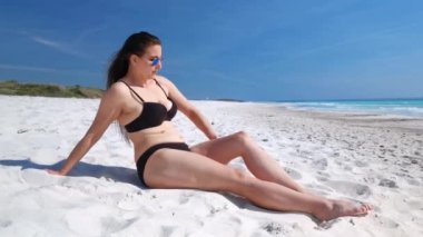 Beyaz bir plajda güneşlenen bir kız bacağındaki kumu temizliyor ve güneş gözlüğünü ayarlıyor.