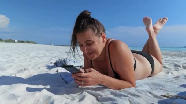 Una Chica Sonriendo Mientras Usa Teléfono Inteligente Acostada Sobre Toalla — Vídeos de Stock