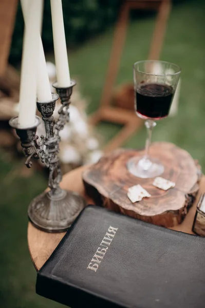 wedding rings, candles, a glass of wine and a bible on the table near wedding arch