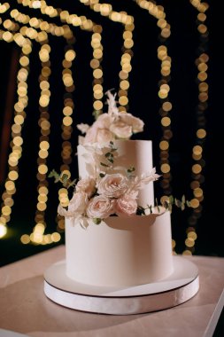 Küçük bir masanın üzerindeki çiçeklerle süslenmiş zarif, iki katlı düğün pastası düğün kemerinin arka planına karşı.