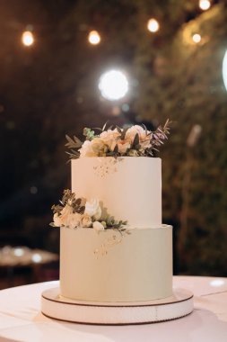 Küçük bir masanın üzerindeki çiçeklerle süslenmiş zarif, iki katlı düğün pastası düğün kemerinin arka planına karşı.