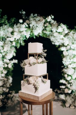 Üç katlı zarif düğün pastası küçük bir masada çiçeklerle süslenmiş düğün kemerinin arka planına karşı.