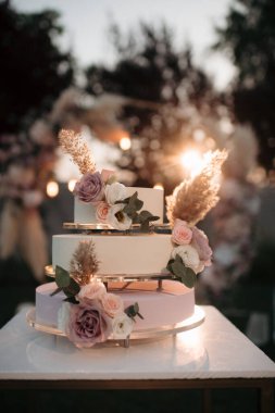 Üç katlı zarif düğün pastası küçük bir masada çiçeklerle süslenmiş düğün kemerinin arka planına karşı.
