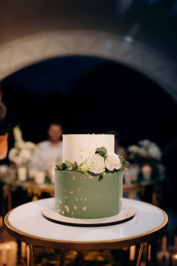 Küçük bir masanın üzerindeki çiçeklerle süslenmiş zarif, iki katlı düğün pastası düğün kemerinin arka planına karşı.