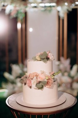 Küçük bir masanın üzerindeki çiçeklerle süslenmiş zarif, iki katlı düğün pastası düğün kemerinin arka planına karşı.