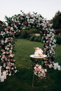 Küçük bir masanın üzerindeki çiçeklerle süslenmiş zarif, iki katlı düğün pastası düğün kemerinin arka planına karşı.