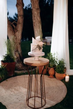 Küçük bir masanın üzerindeki çiçeklerle süslenmiş zarif, iki katlı düğün pastası düğün kemerinin arka planına karşı.