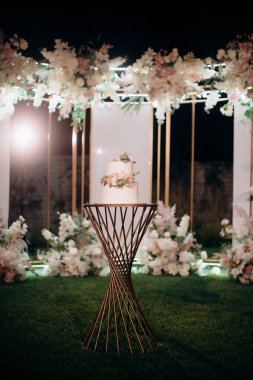 Küçük bir masanın üzerindeki çiçeklerle süslenmiş zarif, iki katlı düğün pastası düğün kemerinin arka planına karşı.