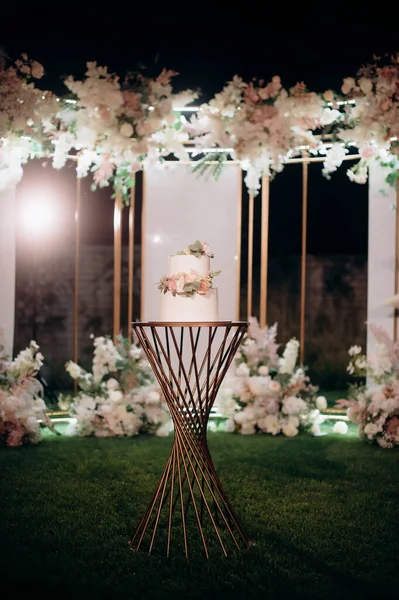 Küçük bir masanın üzerindeki çiçeklerle süslenmiş zarif, iki katlı düğün pastası düğün kemerinin arka planına karşı.