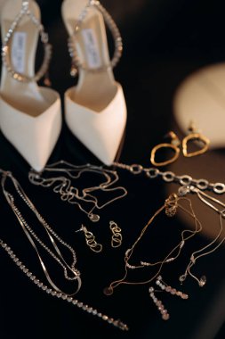 Çekici bir gelin için zarif düğün aksesuarlarının modern bir kompozisyonu. Alyanslar, şık moda ayakkabılar, bir şişe parfüm, siyah arka planda mücevherler.