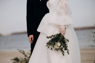 Beyaz şakayık, gül ve okaliptüs demetinin yakın çekimi. Gelin elinde beyaz, zarif bir elbiseyle bir buket tutuyor.