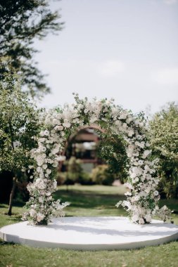 Açık havada düğün töreni. Çimlerde çeşitli taze beyaz çiçekler ve yeşilliklerle süslenmiş büyük, modern bir düğün kemeri.