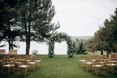 Beyaz çiçeklerin doğal minimalist düğün dekoru ve yeşil çimlerin üzerinde suya bakan ahşap sandalyeler.