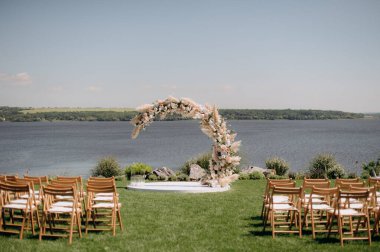 Beyaz çiçeklerin doğal minimalist düğün dekoru ve yeşil çimlerin üzerinde suya bakan ahşap sandalyeler.