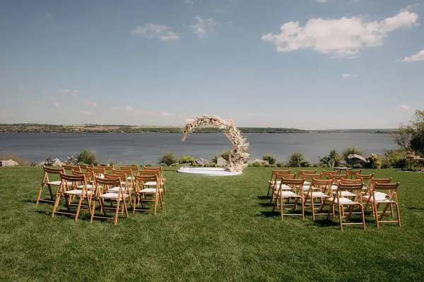 Beyaz çiçeklerin doğal minimalist düğün dekoru ve yeşil çimlerin üzerinde suya bakan ahşap sandalyeler.