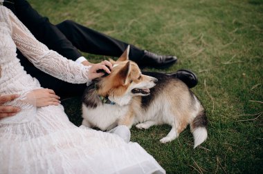 Düğünleri sırasında çimlerin üzerinde oturan gelin ve damadın yanında yatan paslı siyah bir Corgi. Dışarıdaki huzurlu an kutlamaya sıcaklık ve cazibe katar.