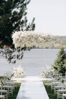 Göl kenarında beyazlarla süslenmiş lüks bir açık hava düğün töreni. Zarif bir çiçek kemeri, şeffaf sandalyeler, yeşil bir çimen ve fevkalade bir arka plan.
