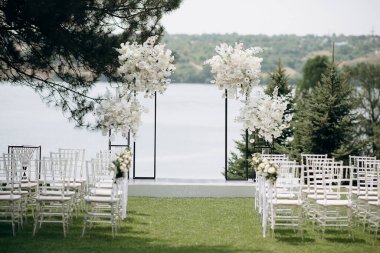 Göl kenarında beyazlarla süslenmiş lüks bir açık hava düğün töreni. Zarif bir çiçek kemeri, şeffaf sandalyeler, yeşil bir çimen ve fevkalade bir arka plan.