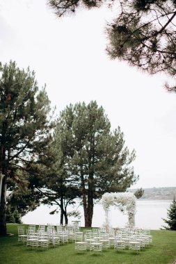Göl kenarında beyazlarla süslenmiş lüks bir açık hava düğün töreni. Zarif bir çiçek kemeri, şeffaf sandalyeler, yeşil bir çimen ve fevkalade bir arka plan.