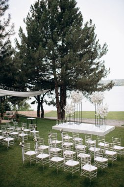 Göl kenarında beyazlarla süslenmiş lüks bir açık hava düğün töreni. Zarif bir çiçek kemeri, şeffaf sandalyeler, yeşil bir çimen ve fevkalade bir arka plan.