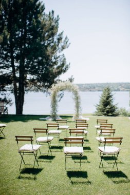 Göl kenarında beyazlarla süslenmiş lüks bir açık hava düğün töreni. Zarif bir çiçek kemeri, şeffaf sandalyeler, yeşil bir çimen ve fevkalade bir arka plan.