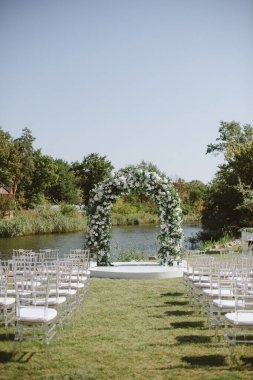 Nehir kenarında çiçek kemeri ve şeffaf sandalyeleri olan güzel bir açık hava düğün töreni. Zarif beyaz ve yeşil dekor doğal ile harmanlanır.