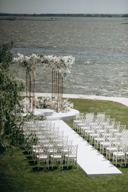 Göl kenarında beyazlarla süslenmiş lüks bir açık hava düğün töreni. Zarif bir çiçek kemeri, şeffaf sandalyeler, yeşil bir çimen ve fevkalade bir arka plan.