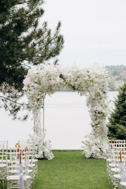 Göl kenarında beyazlarla süslenmiş lüks bir açık hava düğün töreni. Zarif bir çiçek kemeri, şeffaf sandalyeler, yeşil bir çimen, fevkalade doğal bir arka plan.