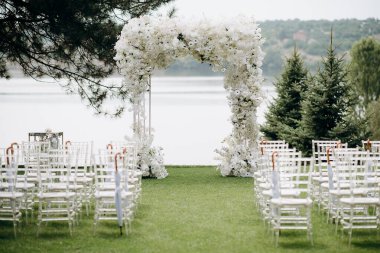 Göl kenarında beyazlarla süslenmiş lüks bir açık hava düğün töreni. Zarif bir çiçek kemeri, şeffaf sandalyeler, yeşil bir çimen, fevkalade doğal bir arka plan.