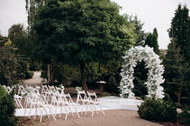 Ağaçlarla çevrili küçük bir avlu, beyaz bir çiçek ve yeşillik kemer, beyaz bir patika, ve açık hava etkinliği için düzenlenmiş beyaz ahşap sandalyeler