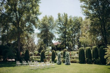 Parkta büyüleyici bir çayır beyaz çiçekler ve yeşilliklerle süslenmiş küçük bir kemer, şeffaf sandalyelerle tamamlanmış sakin bir açık hava düğünü için.