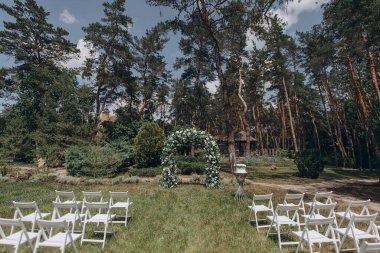 Çam ormanının önündeki bir çayırda düzenlenen düğün töreninde beyaz bir çiçek, yeşil kemer ve zarif beyaz katlanır sandalyeler yer alıyor.