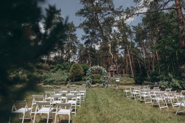 Çam ormanının önündeki bir çayırda düzenlenen düğün töreninde beyaz bir çiçek, yeşil kemer ve zarif beyaz katlanır sandalyeler yer alıyor.
